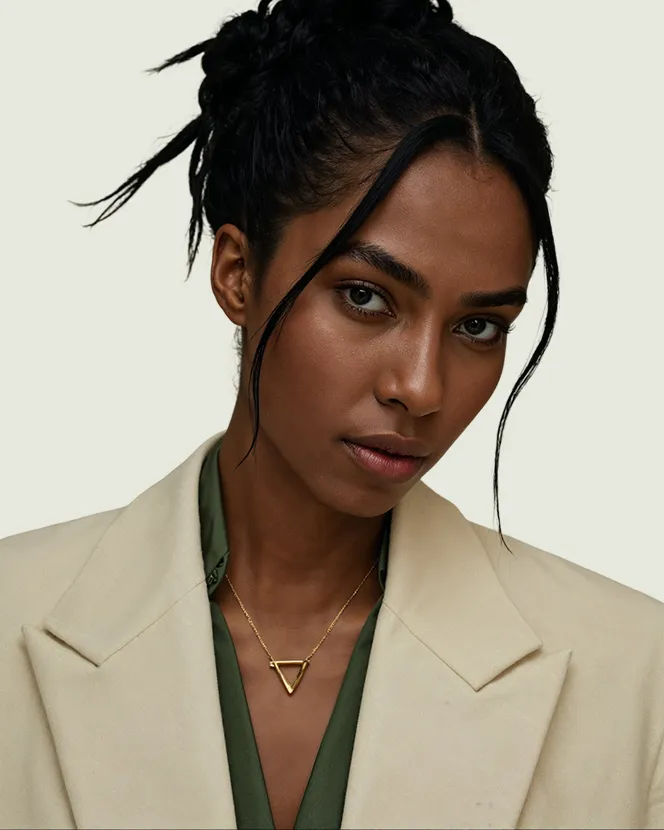 Portrait of a woman with dark braided hair wearing a beige blazer, green blouse, and a gold triangle pendant necklace.